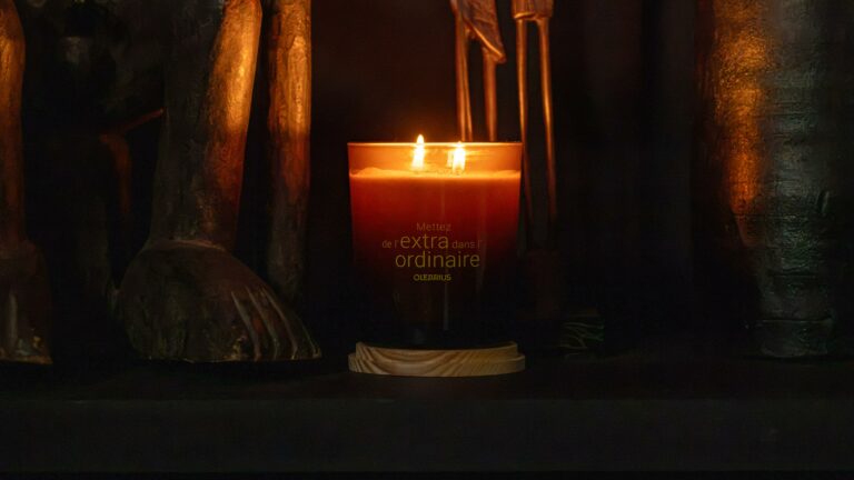 A lit Heritage candle engraved with “Add something extra to the ordinary,” placed between two wooden figurines — lifestyle photography by Olearius · vrai studio