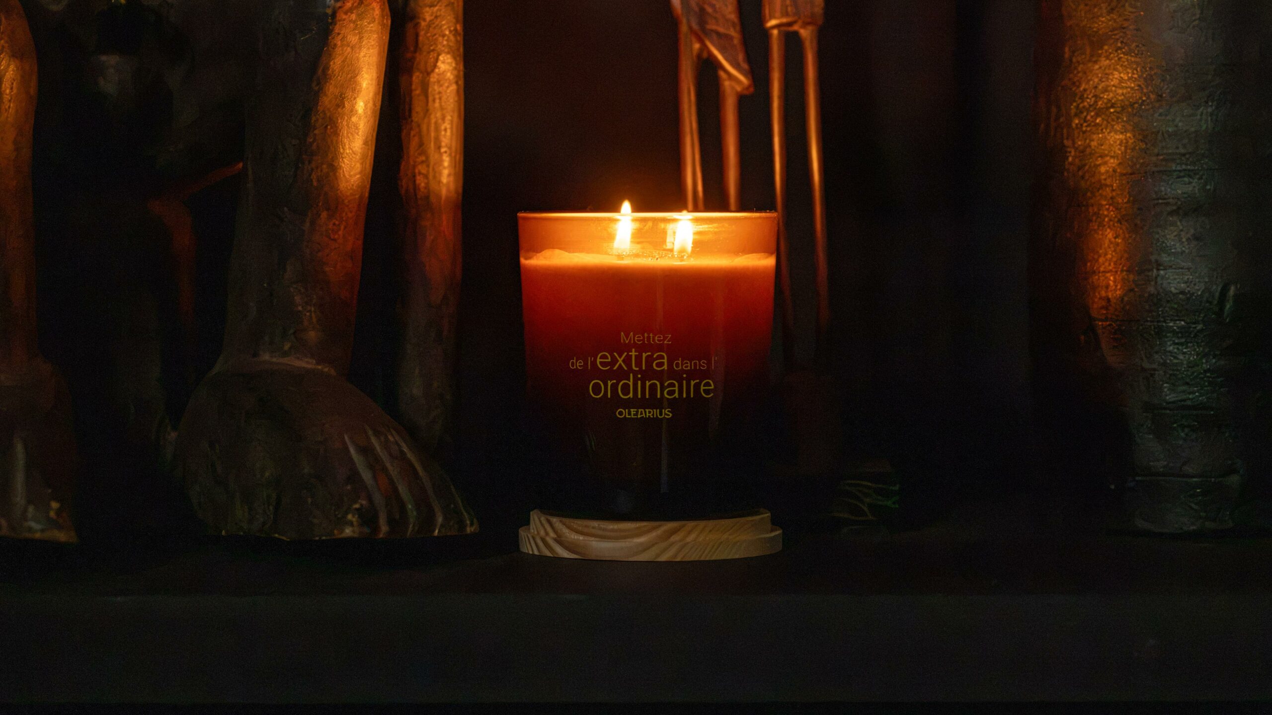 Bougie Heritage allumée gravée « Mettez de l'extra dans l'ordinaire », entre deux statuettes en bois, photographie lifestyle Olearius · vrai studio